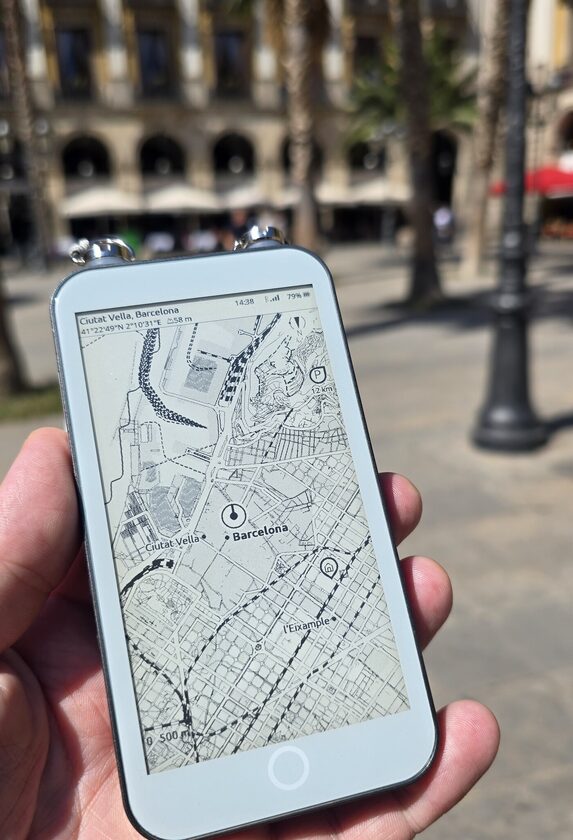 An e-ink reader held flat in one hand in a sunlit Barcelona square, its panel showing an ExplorInk street map of Ciutat Vella with a position dot, a pin at 12 km and a 500 m scale bar, the arcade and palm trees of the square out of focus behind it