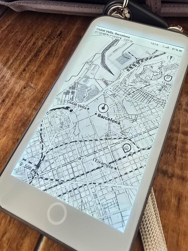 The same e-ink reader lying on a dark wooden cafe table beside a camera bag, its panel filling the frame with an ExplorInk map of central Barcelona in which every street of the Eixample grid is legible