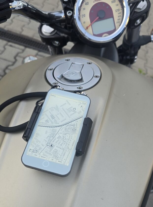 The LilyGo T5 E-Paper S3 Pro held by a black clamp mount on the fuel tank of a parked cruiser, its panel showing an ExplorInk street map with a position marker, pins at the edge and the button labels along the bottom