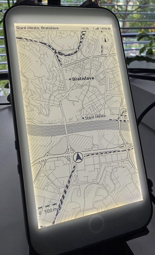 The LilyGo T5 E-Paper S3 Pro standing on a desk with its frontlight on, showing an ExplorInk map of central Bratislava with the Danube across the middle, a position marker, a compass and a scale bar