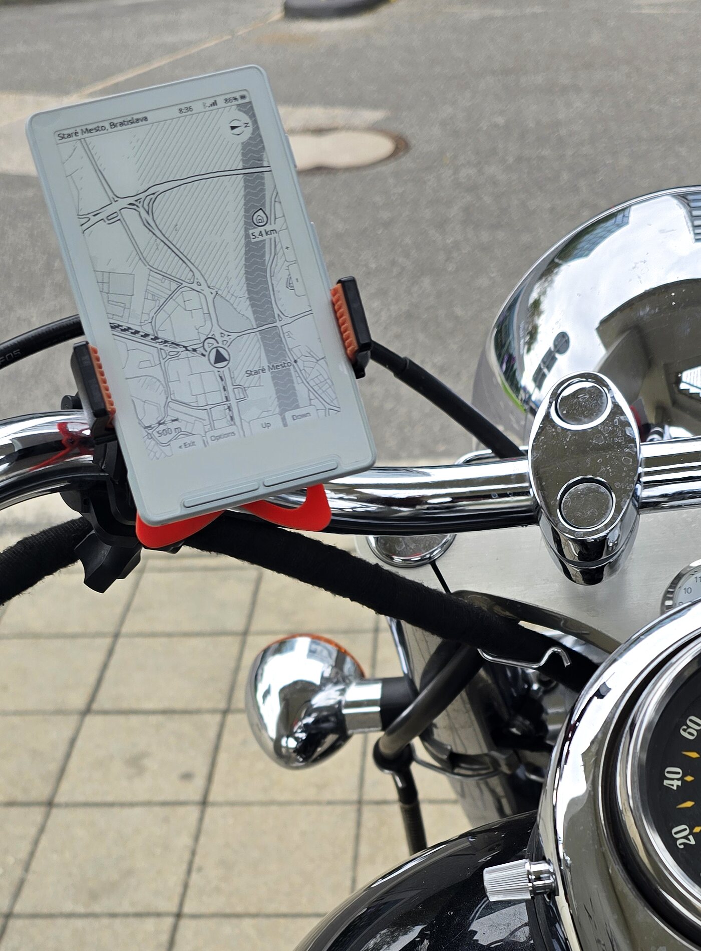Xteink X4 in a bar mount on a motorcycle handlebar, chrome headlight and speedometer beside it, the panel showing a city map with the place name and clock along the top, a home pin at 5.4 km and a 500 m scale bar