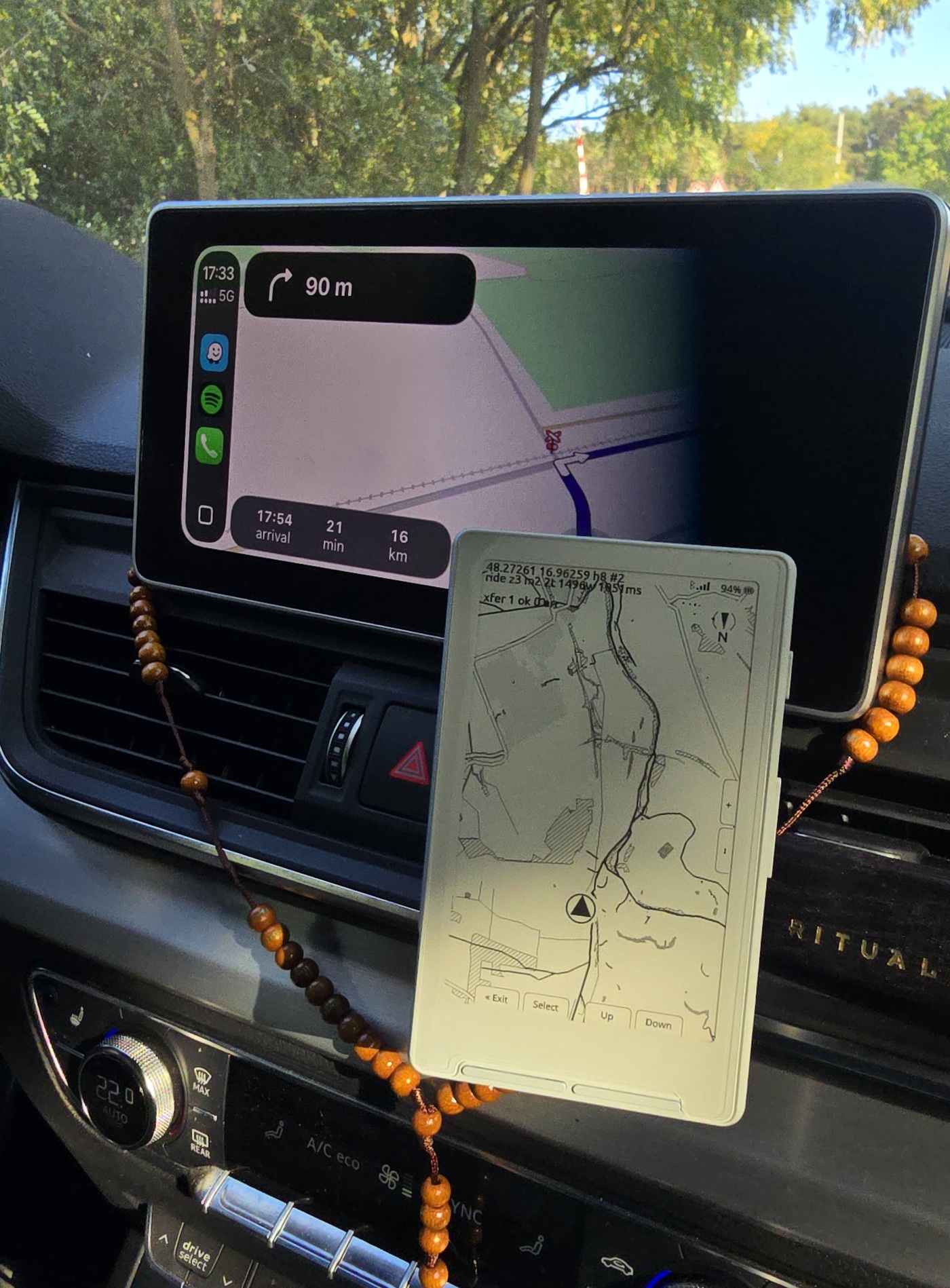 Xteink X4 clipped to a car dashboard mount next to the car's own navigation screen, showing a real schematic map with a position marker, zoom controls and Exit/Select/Up/Down button labels