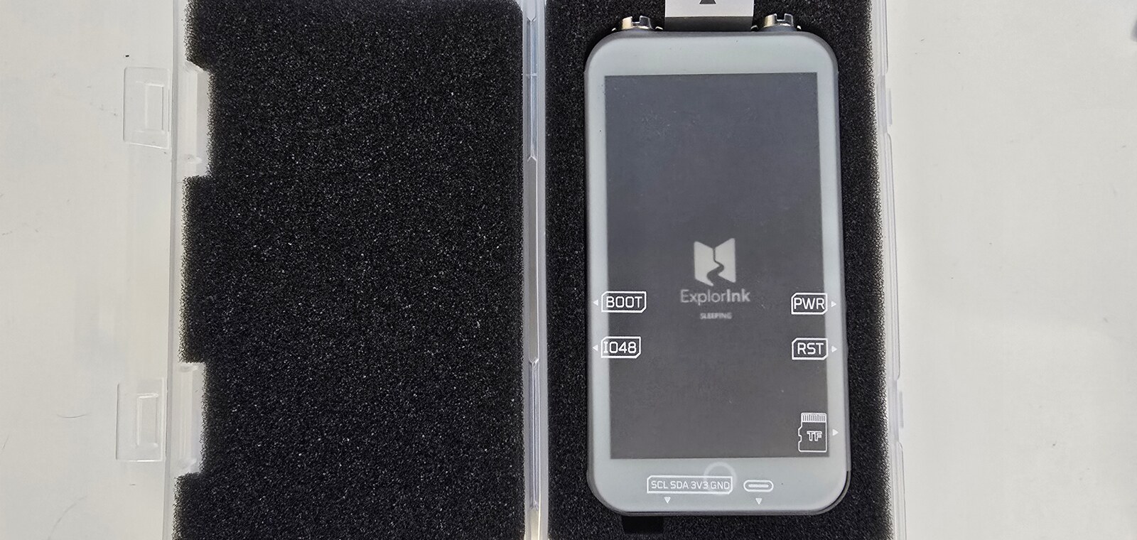The LilyGo T5 E-Paper S3 Pro lying in its open transport case, its panel showing the ExplorInk sleep screen: the logo, the word sleeping, and labels naming the BOOT, IO48, PWR, RST, microSD and SCL SDA 3V3 GND connections along the edges