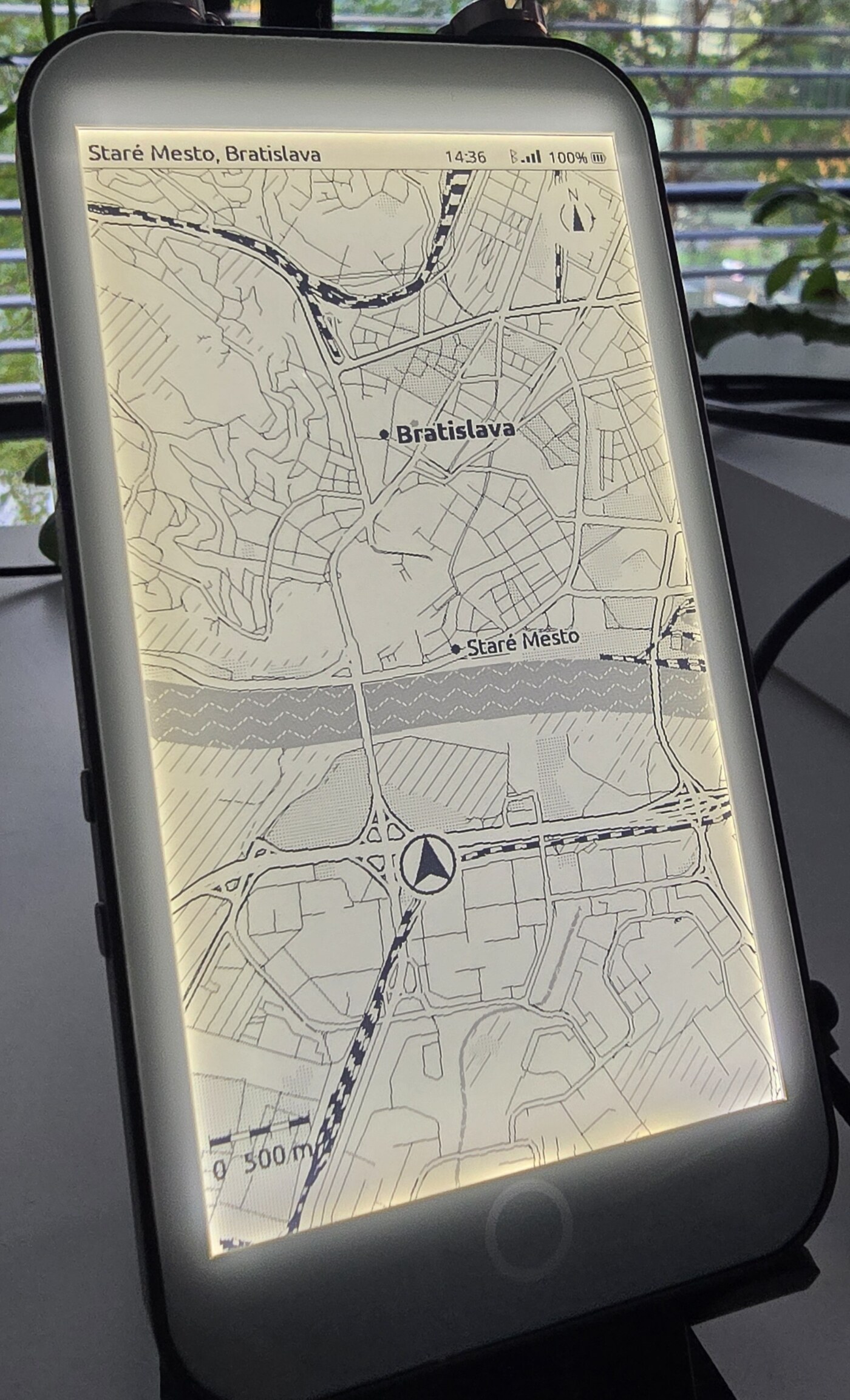 The LilyGo T5 E-Paper S3 Pro standing on a desk, its frontlight on, showing an ExplorInk map of central Bratislava with the Danube across the middle, a position marker, a compass and a scale bar